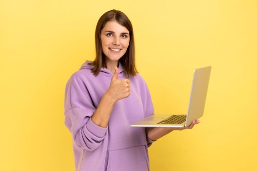 Portrait of optimistic delighted young adult woman working on laptop compute, looking at camera, showing thumb up, wearing purple hoodie. Indoor studio shot isolated on yellow background.