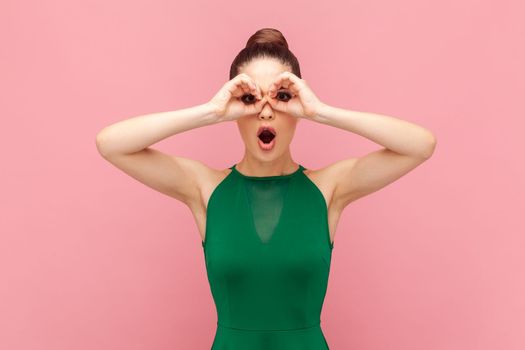 Portrait of funny shocked surprised woman standing with binoculars gesture, looking with open mouth, sees something astonished, wearing green dress. Indoor studio shot isolated on pink background.