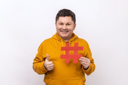 Cheerful delighted man showing thumb up like gesture holding red hashtag sign, satisfied with target advertisement, wearing urban style hoodie. Indoor studio shot isolated on white background.