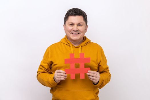 Hashtag and blogging. Portrait of amazing positive man holding big paper hash symbol and smiling at camera, wearing urban style hoodie. Indoor studio shot isolated on white background.