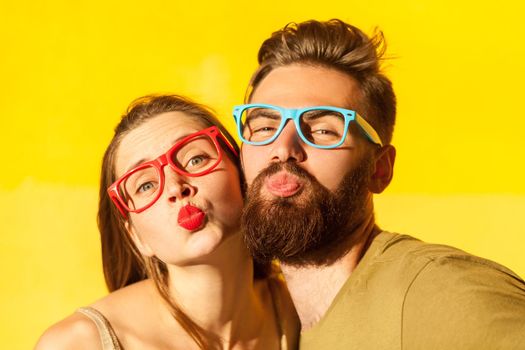 Portrait of romantic pleased loving couple standing looking at camera and sending air kiss, posing with pout lips, being on date. Indoor studio shot isolated on yellow background.