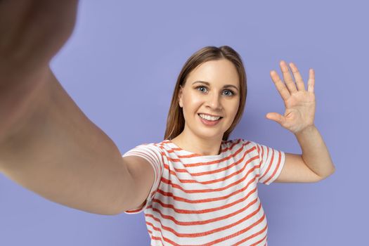 Portrait of attractive woman taking selfie picture, point of view, looking at the camera and waving hand, saying hi, making front selfportrait. Indoor studio shot isolated on purple background.