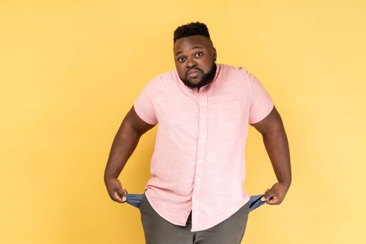 Portrait of frustrated worried man wearing pink shirt turning out empty pockets showing no money gesture, frowning face. Indoor studio shot isolated on yellow background.