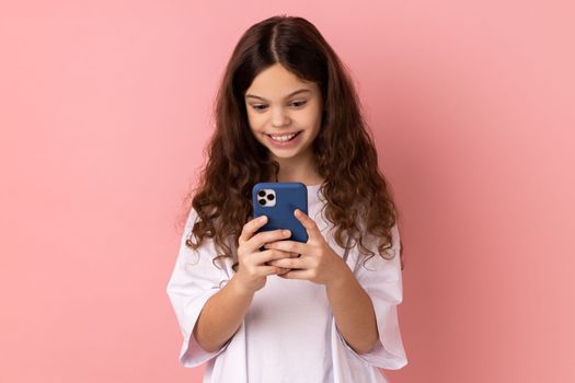 Portrait of happy little girl wearing white T-shirt texting message in social media on cell phone, using mobile network services, chatting online. Indoor studio shot isolated on pink background.