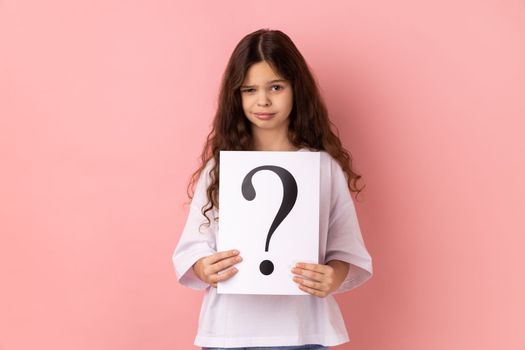 Portrait of winsome beautiful little girl wearing white T-shirt looking at camera, holding paper with question mark, thinks about tasks. Indoor studio shot isolated on pink background.