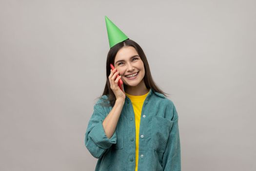 Smiling happy woman in party cone talking phone, celebration her birthday, greeting, congratulating, wearing casual style jacket. Indoor studio shot isolated on gray background.