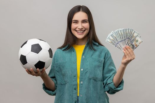 Portrait of smiling woman showing soccer ball and fun of hundred dollar bills, winning lot of money betting for sport, wearing casual style jacket. Indoor studio shot isolated on gray background.