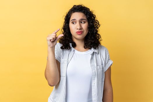 Small amount. Portrait of woman with dark wavy hair showing little bit gesture with hand and looking pleadingly, frowning face. Indoor studio shot isolated on yellow background.