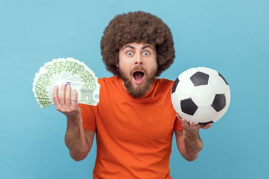 Portrait of surprised man with Afro hairstyle wearing orange T-shirt holding soccer ball and fan of euro bills, sports betting, big win. Indoor studio shot isolated on blue background.