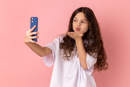 Portrait of little girl wearing white T-shirt sending air kiss on smartphone camera while communicating by video call or streaming vlog. Indoor studio shot isolated on pink background.