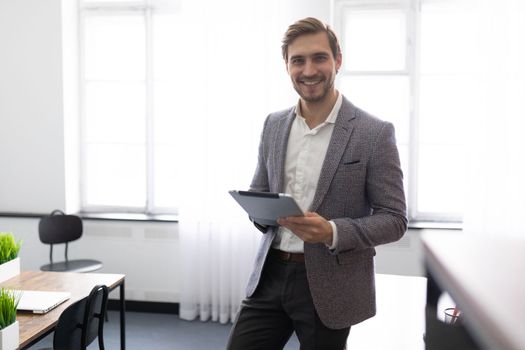 young Businessman with an electronic tablet in his hands stands with a smile in the middle of the office.