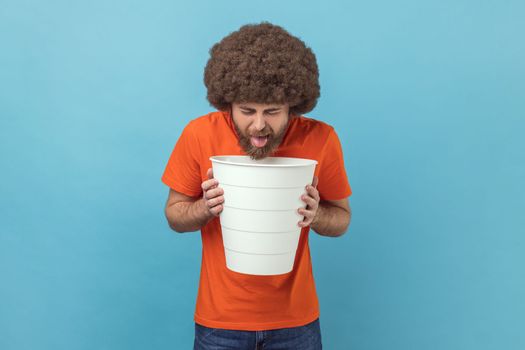 Portrait of man with Afro hairstyle wearing orange T-shirt suffering from bad feelings, discomfort in stomach, being sick, spoiled product. Indoor studio shot isolated on blue background.