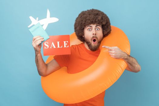 Shocked man with Afro hairstyle standing with orange rubber ring, holding passport document and pointing sale card, tours for low cost. Indoor studio shot isolated on blue background