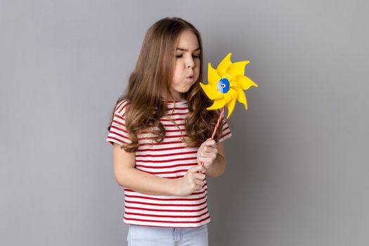 Origami hand mill. Portrait of satisfied little girl wearing striped T-shirt blowing at paper windmill, playing with pinwheel toy on stick. Indoor studio shot isolated on gray background.