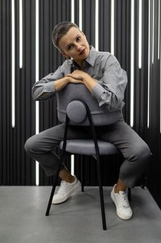 a stylish woman of European appearance sits on a chair in a gray shirt against a gray wall.