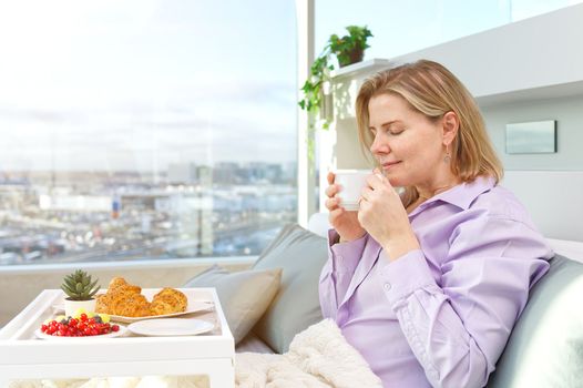 Happy middle aged, beautiful woman having breakfast, relaxing in hotel bed. Woman having breakfast sitting on her bed in luxury apartments. enjoying lazy morning lifestyle