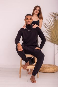 A happy beautiful multiracial couple, in black clothes, are sitting near the wall in the room, smiling, look at camera.Vertical. Copy space