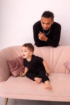 Happy father and little son, in black clothes, sitting on ligh pink sofa in the room, light interior. Vertical. Copy space