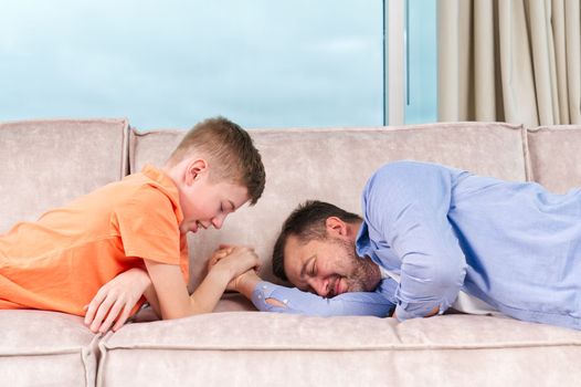 Handsome young father and his cute son competing in arm wrestling while lying on a sofa. Father and son quality time