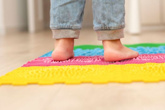 Health concept. A little boy stands with bare feet on a multi-colored orthopedic massage mat in a home interior. Close-up of children's heels. Back view.