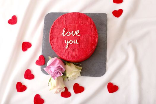 Small delicious red bento cake with the inscription Love you, with red hearts and roses lying on a white bed next to it. Top view. Close up. Copy space