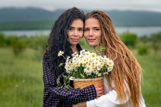 Two women enjoy nature in a field of daisies. Girlfriends hugging hold a bouquet of daisies and look at the camera