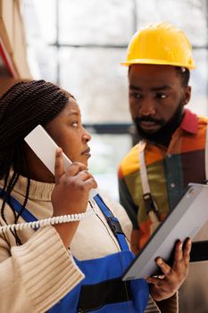African american woman holding tablet while talking on landline phone in warehouse. Logistics manager consulting with storehouse worker and answering supervisor call in storage room