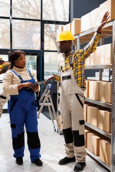 African american warehouse worker explaining supervisor packages storage system, showing on parcels shelf and talking. Storehouse manager listening to colleague and holding digital tablet