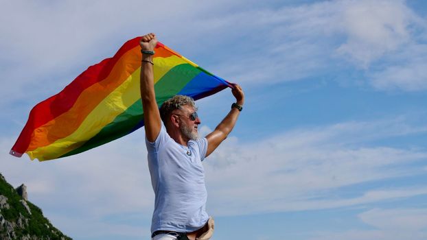 Portrait of a gray-haired elderly Caucasian man with a beard and sunglasses holding a rainbow LGBTQIA flag against a sky background. Bisexual Gay Celebrates Pride Month Coming Out Day