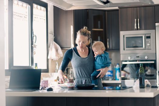 Happy mother and little infant baby boy together making pancakes for breakfast in domestic kitchen. Family, lifestyle, domestic life, food, healthy eating and people concept