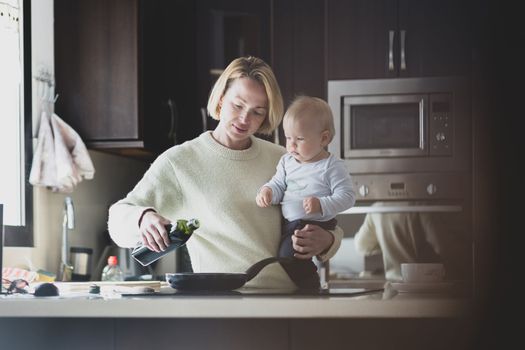 Happy mother and little infant baby boy together making pancakes for breakfast in domestic kitchen. Family, lifestyle, domestic life, food, healthy eating and people concept