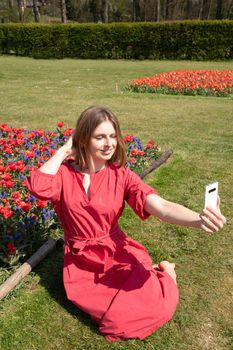 A young beautiful girl in a red dress in a spring park plays with her hair, flirts and takes a selfie on a mobile phone, tulips bloom and people walk in the park, pamper themselves.High quality photo