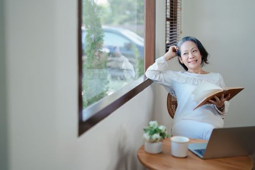 Portrait of an elderly Asian woman in a modern pose holding a memory notebook and operating a computer