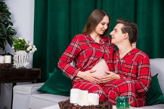 A pair of brides look deeply into each other's eyes while sitting on a gray sofa. A green curtain can be seen in the background. The girl sits on the boy's lap and her pregnant belly is exposed