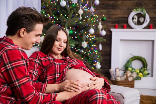 A couple expecting a baby sits close together on a couch. In the background a dressed Christmas tree with colorful lights. Smiling parents-to-be look at the pregnant belly.