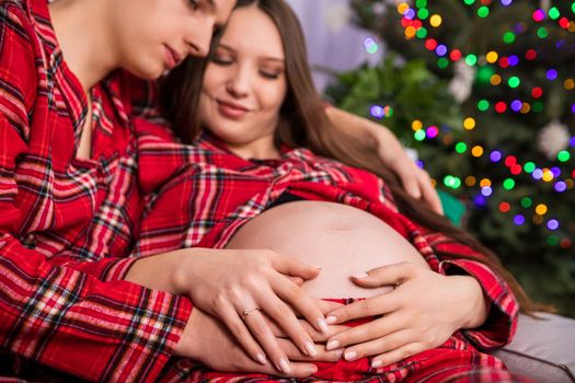 Parents-to-be hold their hands on a woman's pregnant belly. A diamond ring on the woman's hand, a fuzzy Christmas tree with lights in the background.