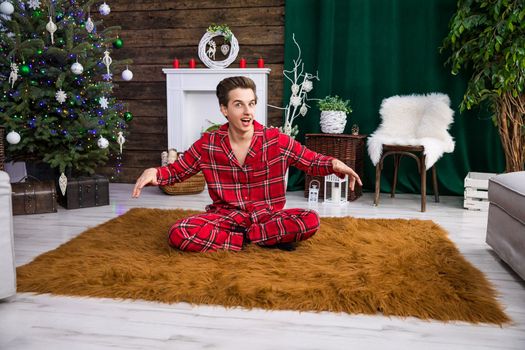 A boy sits on a fluffy brown carpet with his legs crossed and his arms slightly raised. In the background stands a dressed Christmas tree with colorful lights.