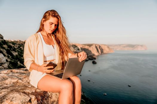 Successful business woman in yellow hat working on laptop by the sea. Pretty lady typing on computer at summer day outdoors. Freelance, travel and holidays concept.