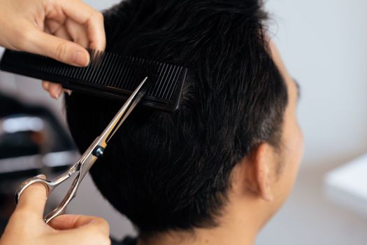 Closeup asian female barber making haircut for male customer with male hairstyle in qualified barbershop. Men's hairstyling by a scissor and comb in hair salon concept.