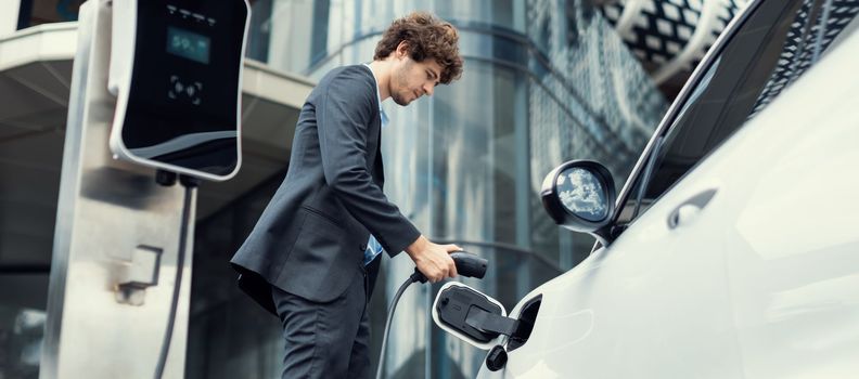 Below view closeup image of progressive black suit businessman recharge battery of his electric vehicle from public charging station. Renewable and alternative energy powered car concept.