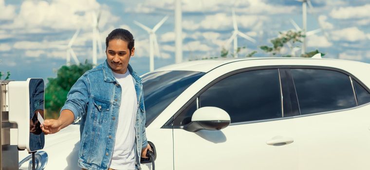 Progressive man with his electric car, EV car recharging energy from charging station on green field with wind turbine as concept of future sustainable energy. Electric vehicle with energy generator.