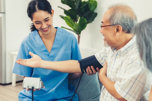 An elderly man having a blood pressure check by his personal caregiver with his wife sitting next to him in their home.