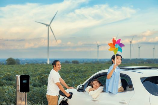 Concept of progressive happy family enjoying their time at wind farm with electric vehicle. Electric vehicle driven by clean renewable energy from wind turbine generator for charging station.