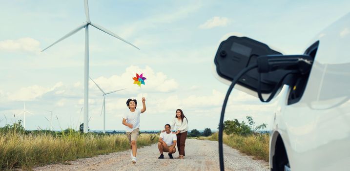 Concept of progressive happy family holding windmill toy and relax at wind farm with electric vehicle. Electric vehicle driven by clean renewable energy from wind turbine generator to charger station.