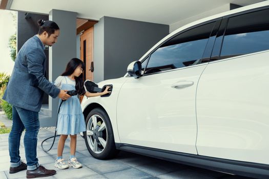 Progressive father and daughter plugs EV charger from home charging station to electric vehicle. Future eco-friendly car with EV cars powered by renewable source of clean energy.