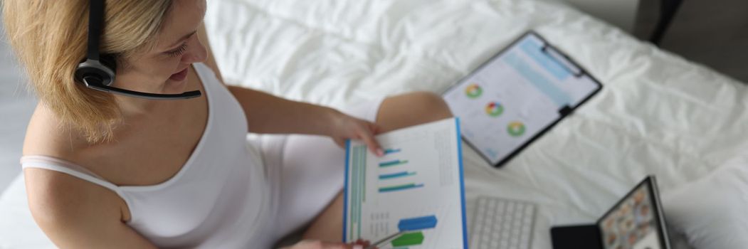 Woman in headphones holds charts sits on bed and holds online conference. Remote work and freelancer