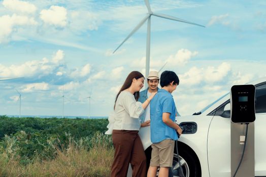 Concept of progressive happy family enjoying their time at wind farm with electric vehicle. Electric vehicle driven by clean renewable energy from wind turbine generator for charging station.