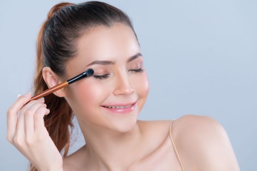 Closeup beautiful girl with flawless applying glamorous eye shadow makeup with eyeliner brush. Cosmetic facial painting process on lovely young woman with perfect clean skin in isolated background.