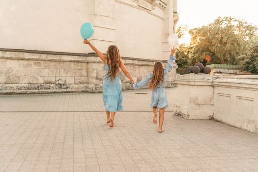 Daughter mother run holding hands. In blue dresses with flowing long hair, they hold balloons in their hands against the backdrop of a sunset and a white building