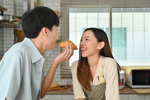 Romantic young couple eating croissant while having breakfast in kitchen, enjoying leisure weekend time together.